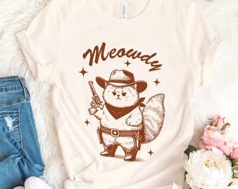 May include: A beige t-shirt with a brown illustration of a cat wearing a cowboy hat and holding a gun. The text "Meowdy" is above the cat.