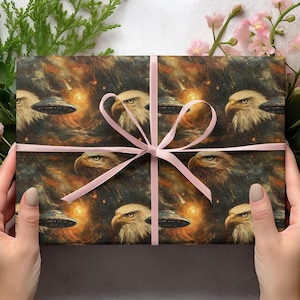 May include: Gift wrap featuring a repeating pattern of bald eagles and UFOs against a cosmic background of orange, brown, and black. A light pink ribbon is tied around the gift. The overall theme is patriotic and space-themed.