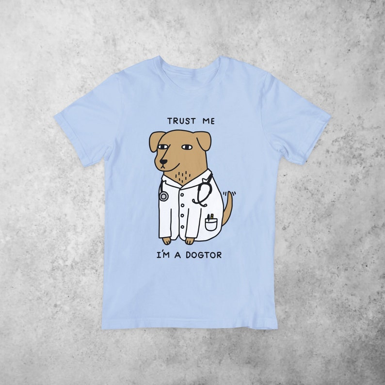Puede incluir: Camiseta azul claro con un perro de dibujos animados que lleva una bata de m&eacute;dico blanca y un estetoscopio. El texto "TRUST ME I'M A DOGTOR" est&aacute; impreso en la camiseta.