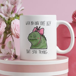 Op de afbeelding: Een witte keramische mok met een groene leguaan die een roze strik draagt en de tekst "When you know you're ugly but still trying..."