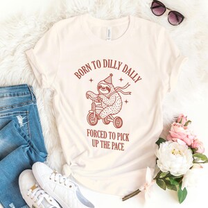 May include: A white t-shirt with a brown graphic of a sloth riding a tricycle. The text "Born to Dilly Dally Forced to Pick Up the Pace" is printed on the shirt.