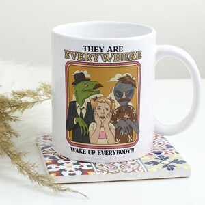 May include: White ceramic mug with a retro-style graphic of a woman, a dinosaur, and an alien. The text on the mug reads "They are Everywhere. Wake up everybody!!"