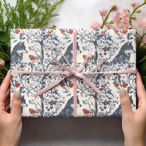 Op de afbeelding: Wit cadeaupapier met een herhalend patroon van blauwe wolven, rode bessen en blauwe en witte bloemenmotieven. Het cadeau is vastgebonden met een roze lint.