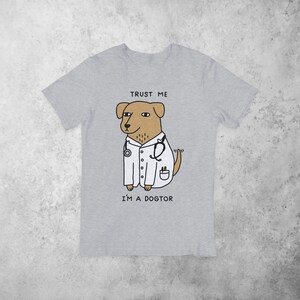 Puede incluir: Camiseta gris con un perro de dibujos animados que lleva una bata de laboratorio blanca y un estetoscopio. El texto "Trust me I'm a dogtor" est&aacute; impreso en la camiseta.