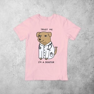 Puede incluir: Camiseta rosa con un perro de dibujos animados que lleva una bata de m&eacute;dico blanca y un estetoscopio. El texto "Trust me I'm a dogtor" est&aacute; impreso en la camiseta.