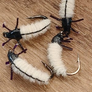 Può includere: Quattro mosche da pesca artificiali. Ogni mosca presenta un corpo bianco e soffice con una striscia nera, una testa nera e zampe viola scuro. Le mosche sono attaccate a ami d'argento curvi. Lo sfondo è una superficie di legno marrone chiaro.