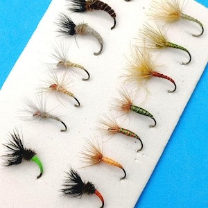Puede incluir: Una colección de moscas de pesca dispuestas sobre una superficie blanca sobre un fondo azul. Las moscas presentan varios colores, incluyendo negro, marrón, beige y verde, con plumas detalladas y anzuelos curvos. Las moscas están diseñadas para la pesca.