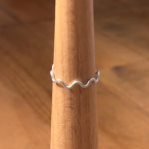 May include: A silver ring featuring a wavy pattern. The ring is shown on a wooden ring display. The design of the ring mimics a sequence of connected waves, offering a distinctive and fashionable appearance. The ring is a simple yet elegant piece of jewellery.