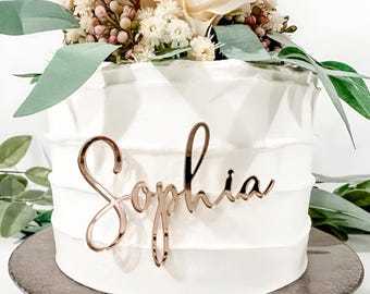 Può includere: Una torta bianca con un topper di rosa e verde. La torta è decorata con un topper in corsivo che dice "Sophia".