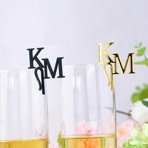 May include: Two champagne flutes with gold-colored liquid are adorned with initial letter charms. One charm is black, the other is gold. The charms feature the letters "K" and "M", likely for a wedding or special event.