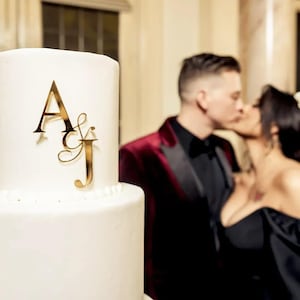 May include: A white wedding cake with gold lettering that says "A & J" on the top tier. A couple is kissing in the background.