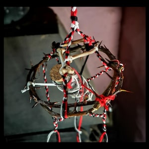 May include: A gothic-style decorative piece. A glass bottle with a cork stopper is suspended within a thorny wreath. The wreath is decorated with bone fragments and wrapped with red, black, and white twine, creating a unique aesthetic.