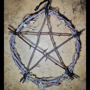 Puede incluir: Un pentagrama rústico elaborado con ramas espinosas y ramitas, atadas con hilo negro. El diseño en forma de estrella presenta un punto central y está adornado con pequeñas hojas, creando una estética natural y terrosa. Se adjunta un lazo de hilo negro para colgar.