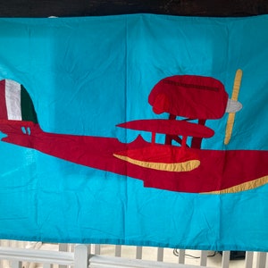 Peut inclure: Une tenture murale en tissu turquoise représentant un avion rouge. L'avion a une hélice jaune, une queue blanche, verte et rouge, et un ventre jaune. L'avion est posé sur un fond turquoise uni.