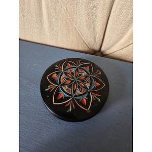May include: A round, black wooden box with a colorful floral design on the lid. The design features red, blue, yellow, and green accents. The box is sitting on a blue surface.