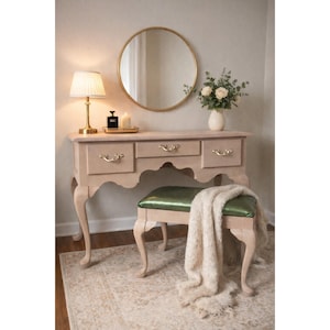 Puede incluir: Una cómoda de color beige claro con tres cajones y tiradores ornamentados, combinada con un taburete a juego con un cojín verde. Una manta esponjosa de color crema descansa sobre el taburete. Un espejo con marco dorado cuelga encima.