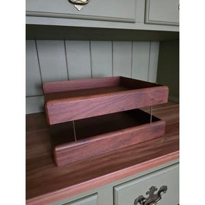 Könnte beinhalten: Ein zweistufiges Holztablett mit rotbraunem Finish. Das Tablett wird von dünnen, goldfarbenen Metallstäben getragen. Der Hintergrund zeigt einen grünen Schrank mit dekorativen Beschlägen.