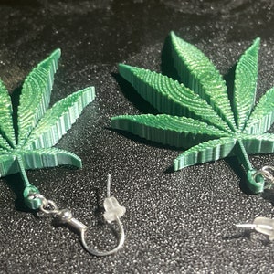 Op de afbeelding: Een paar groene marihuana-bladeren oorbellen met zilveren haken. De bladeren zijn gedetailleerd en hebben een 3D-effect.