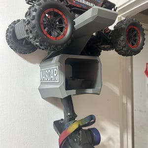 May include: A red and black remote control truck mounted on a grey wall-mounted holder. The holder has a compartment with the word "NASCAR" on it. A remote control with a colourful strap is attached to the holder.