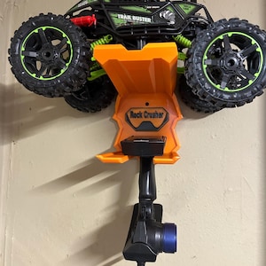 May include: A black and green Trail Buster remote control truck with large, rugged tires and neon green accents. The orange Rock Crusher base holds the remote control, which has a blue end.