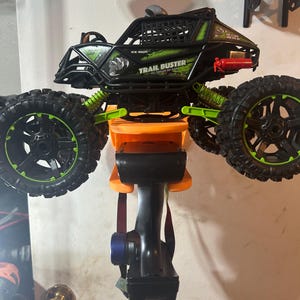 May include: A black and green remote control toy truck with a black and green remote control. The truck is labeled "Trail Buster" and has a black cage on the back. The truck is mounted on an orange wall mount.