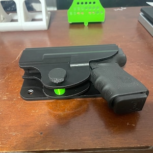 May include: A black gun holster with a green adjustment knob, mounted on a wooden surface. The holster is designed to hold a black handgun with a black magazine.