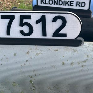 May include: A gray metal mailbox with a black and white oval sign that reads "7512".