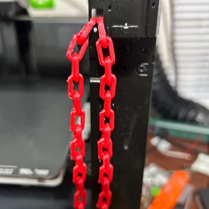 May include: Two red plastic chains hanging from a black metal object. The chains are made of interlocking links.