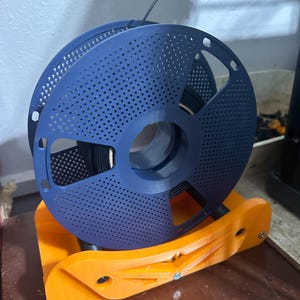 Könnte beinhalten: Eine blaue 3D-Drucker-Filamentspule mit perforiertem Design, montiert auf einem orangefarbenen Spulenhalter. Das Filament ist dunkel und der Halter besteht aus Kunststoff. Das Setup ist für den 3D-Druck konzipiert.