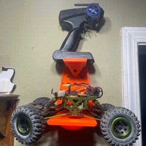 Op de afbeelding: Oranje wandbevestiging voor een radiografisch bestuurbare auto en een afstandsbediening. De auto is groen en zwart en heeft vier wielen. De afstandsbediening is zwart en heeft een blauwe antenne.