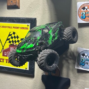 May include: A green and black monster truck with large tires is mounted on a wall. The truck is displayed next to a sign that reads "HE 251 BARNYARD 2019 FALL POINT SERIES 2ND PLACE 4WD SCT TR". There are two stickers on the wall, one that reads "B&T RC RACEWAY 3rd" and another that reads "RC RACEWAY 2nd".