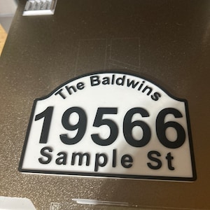 May include: White and black 3D printed house number sign with the text "The Baldwins" and "19566 Sample St."