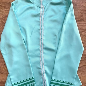 May include: A light turquoise jacket with a high collar and long sleeves. The jacket has white lace trim along the zipper and collar, with green leaf and dot patterns on the cuffs. The fabric has a slight sheen.