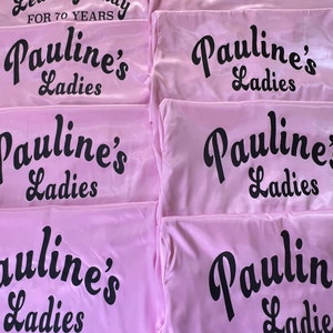 May include: Pink satin robes with black text that reads "Leading Lady for 70 Years" and "Pauline's Ladies". The robes are folded and stacked, showcasing the text prominently. The image is well-lit.