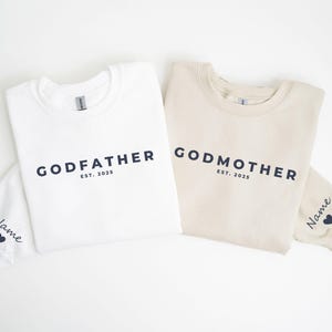 May include: Two crewneck sweatshirts, one white and one beige, with the words "GODFATHER" and "GODMOTHER" printed in navy blue. Each sweatshirt has "EST. 2025" printed below the main text. Sleeves have a name and heart.