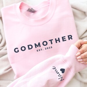 Pu&ograve; includere: Una felpa rosa chiaro con la scritta "GODMOTHER" in blu navy, insieme a "EST. 2025". La manica mostra un nome e un simbolo a forma di cuore. La felpa &egrave; piegata su una coperta colorata neutra.