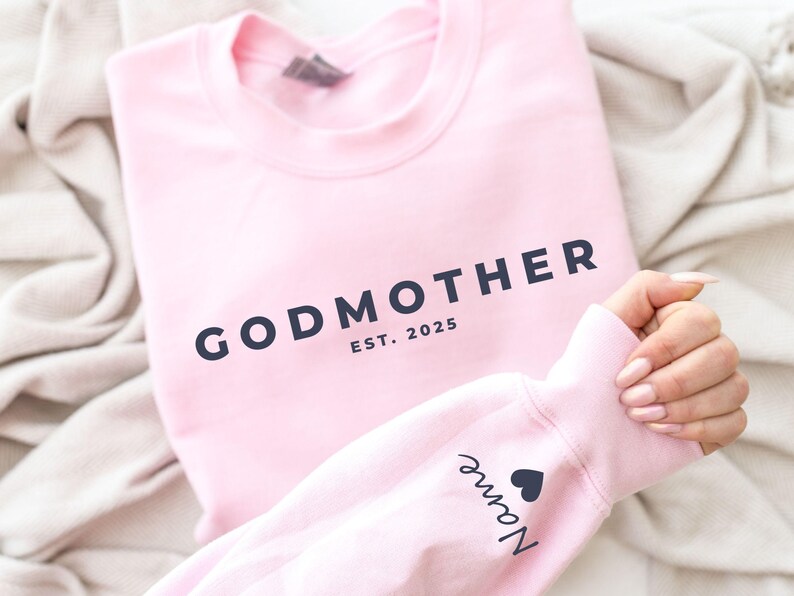 Pu&ograve; includere: Una felpa rosa chiaro con la scritta "GODMOTHER" in blu navy, insieme a "EST. 2025". La manica mostra un nome e un simbolo a forma di cuore. La felpa &egrave; piegata su una coperta colorata neutra.