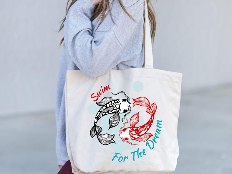 May include: Cream-colored tote bag featuring a black and white koi fish and a red koi fish in a yin-yang design. The text "Swim For The Dream" is printed in red and teal. The bag has a simple, minimalist design.
