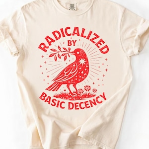 May include: Cream-coloured t-shirt with a red graphic. The graphic features a red bird holding a branch, surrounded by the words "RADICALIZED BY BASIC DECENCY" in a circular design. The shirt has short sleeves and a crew neck.