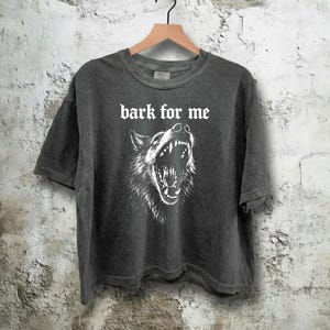 May include: Dark gray t-shirt with the text "bark for me" above a detailed white illustration of a wolf's snarling face. The shirt is hanging on a wooden hanger against a textured wall.