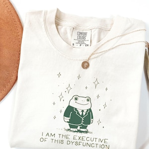 Puede incluir: Camiseta color crema con una rana de dibujos animados con traje, rodeada de estrellas. El texto "I AM THE EXECUTIVE OF THIS DYSFUNCTION" está impreso debajo. Un collar dorado con un colgante está sobre la camiseta.
