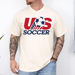 Puede incluir: Una camiseta de manga corta de color crema con un dise&ntilde;o gr&aacute;fico que presenta el texto rojo US, un bal&oacute;n de f&uacute;tbol azul y blanco que sustituye a la letra S, y la palabra SOCCER en azul debajo. El dise&ntilde;o incluye una franja azul con estrellas.