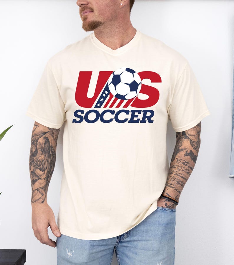 Puede incluir: Una camiseta de manga corta de color crema con un dise&ntilde;o gr&aacute;fico que presenta el texto rojo US, un bal&oacute;n de f&uacute;tbol azul y blanco que sustituye a la letra S, y la palabra SOCCER en azul debajo. El dise&ntilde;o incluye una franja azul con estrellas.
