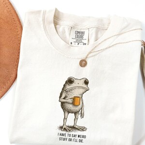 May include: Cream-coloured Comfort Colors t-shirt with a cartoon frog holding a mug. The frog has large eyes and a serious expression. The text "I have to say weird stuff or I'll die" is printed below the frog. A gold necklace and a brown hat are also visible.