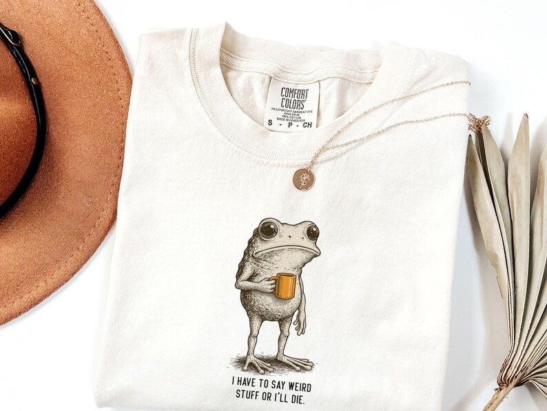 May include: Cream-coloured Comfort Colors t-shirt with a cartoon frog holding a mug. The frog has large eyes and a serious expression. The text "I have to say weird stuff or I'll die" is printed below the frog. A gold necklace and a brown hat are also visible.