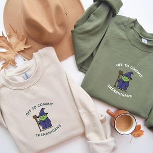 以下が含まれることがあります： 「OFF TO COMMIT SHENANIGANS」の文字とカエルの魔法使いの漫画が描かれた、クリーム色とオリーブグリーンの2つのクルーネックのスウェットシャツ。茶色の帽子、秋の葉、コーヒーカップも画像にあります。