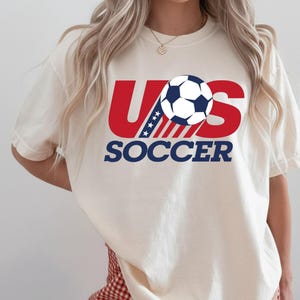 Puede incluir: Una camiseta de manga corta color crema con un dise&ntilde;o gr&aacute;fico que presenta el texto rojo US, un bal&oacute;n de f&uacute;tbol azul y blanco que reemplaza la letra O, y la palabra SOCCER en azul debajo. La letra U incluye una secci&oacute;n azul con estrellas blancas.