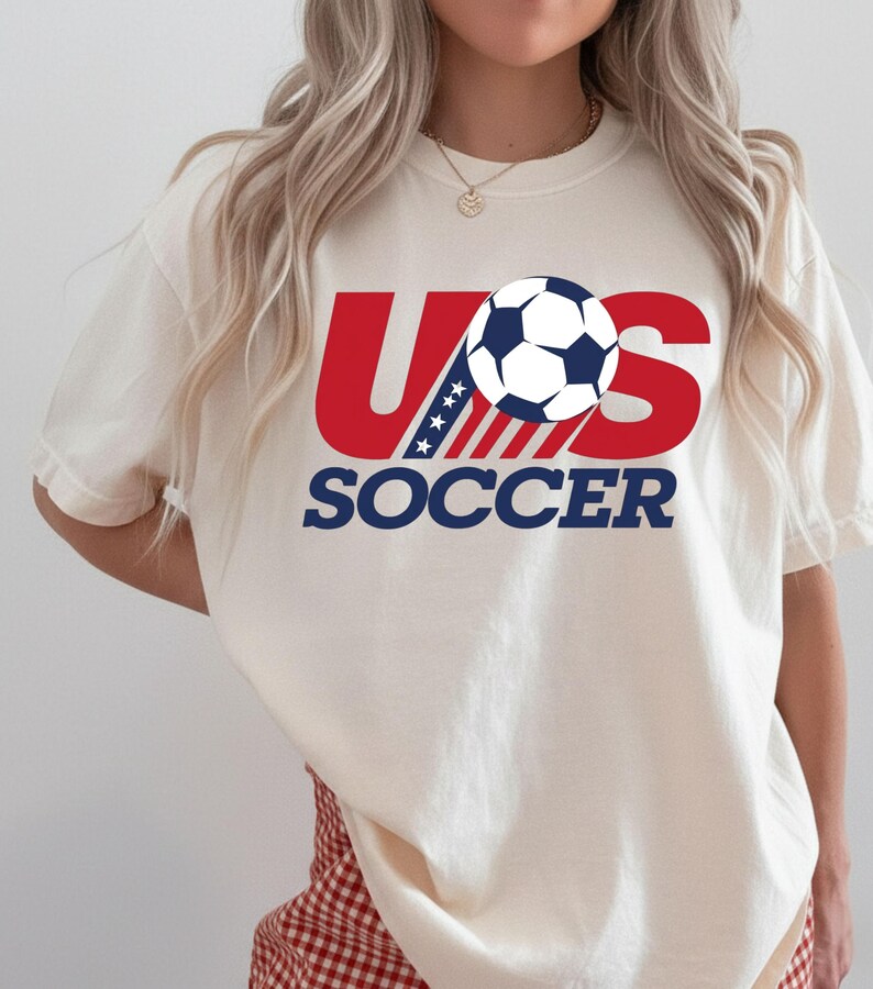 Puede incluir: Una camiseta de manga corta color crema con un dise&ntilde;o gr&aacute;fico que presenta el texto rojo US, un bal&oacute;n de f&uacute;tbol azul y blanco que reemplaza la letra O, y la palabra SOCCER en azul debajo. La letra U incluye una secci&oacute;n azul con estrellas blancas.