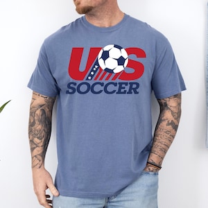 Puede incluir: Una camiseta azul de manga corta con un dise&ntilde;o gr&aacute;fico que presenta el texto rojo US, un bal&oacute;n de f&uacute;tbol que sustituye a la letra O y la palabra SOCCER en azul oscuro debajo. El dise&ntilde;o incluye una franja diagonal con estrellas blancas.