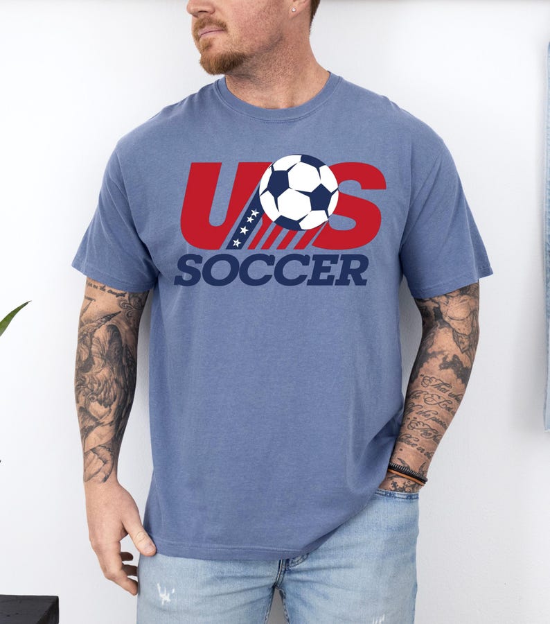Puede incluir: Una camiseta azul de manga corta con un dise&ntilde;o gr&aacute;fico que presenta el texto rojo US, un bal&oacute;n de f&uacute;tbol que sustituye a la letra O y la palabra SOCCER en azul oscuro debajo. El dise&ntilde;o incluye una franja diagonal con estrellas blancas.